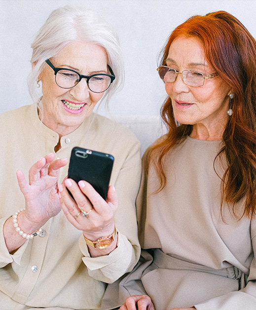 Zwei lächelnde ältere Damen mit Brille, die gemeinsam auf einem weißen Sofa sitzen und per Smartphone etwas online bestellen.